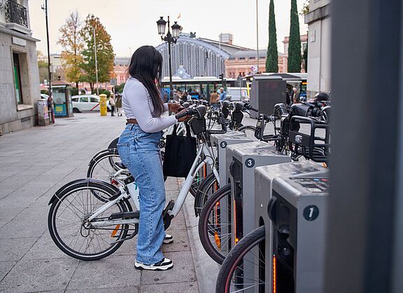 Eine junge Frau mit langen dunklen Haaren, weißem Oberteil, Jeans und schwarzer Umhängetasche steht an einer Bike-Sharing-Station und entnimmt ein weißes Leihfahrrad. Die Station verfügt über mehrere nummerierte Stellplätze mit grauen Dockingelementen. Im Hintergrund sind ein belebter Stadtplatz, historische Gebäude, Passanten und ein Bahnhofsgebäude zu sehen.